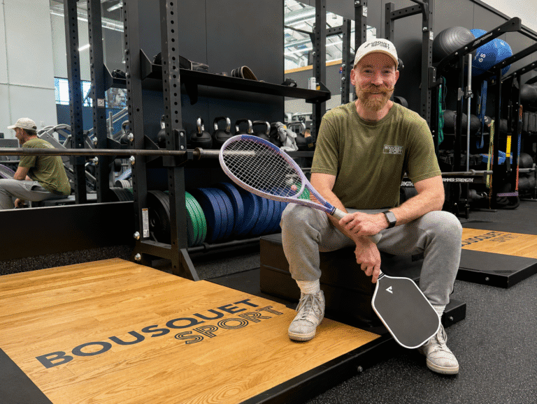 A smiling man in a green t-shirt sits on a gym plyo box, holding a purple tennis racket and a training pad in a weight room.