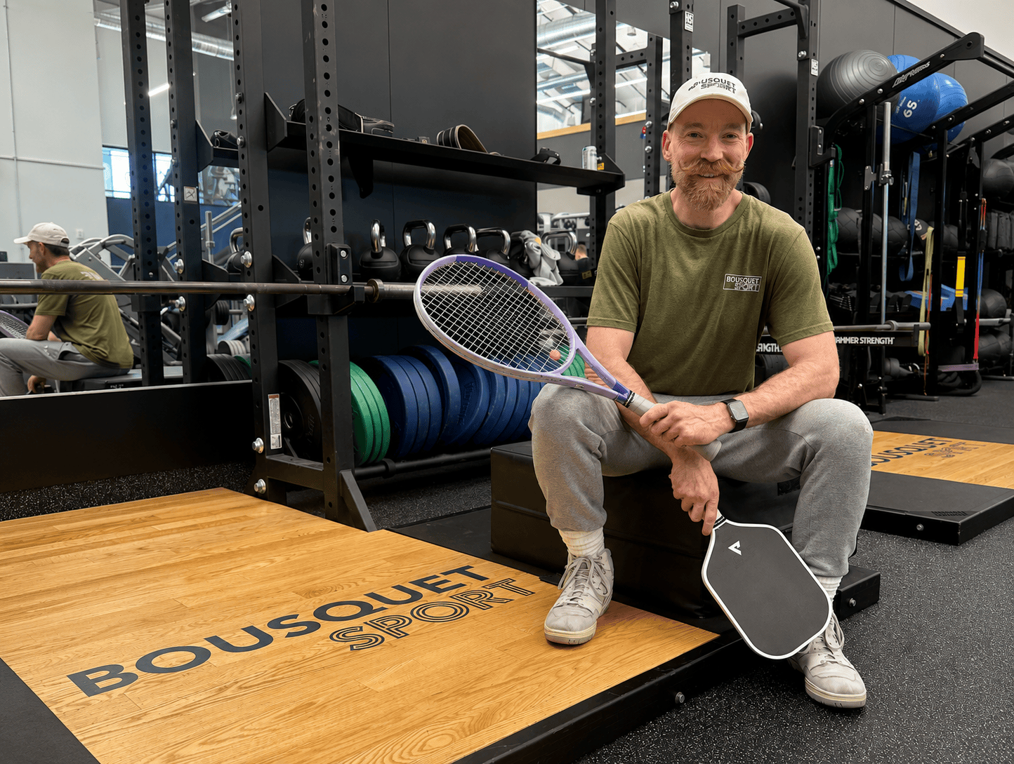 A smiling man in a green t-shirt sits on a gym plyo box, holding a purple tennis racket and a training pad in a weight room.