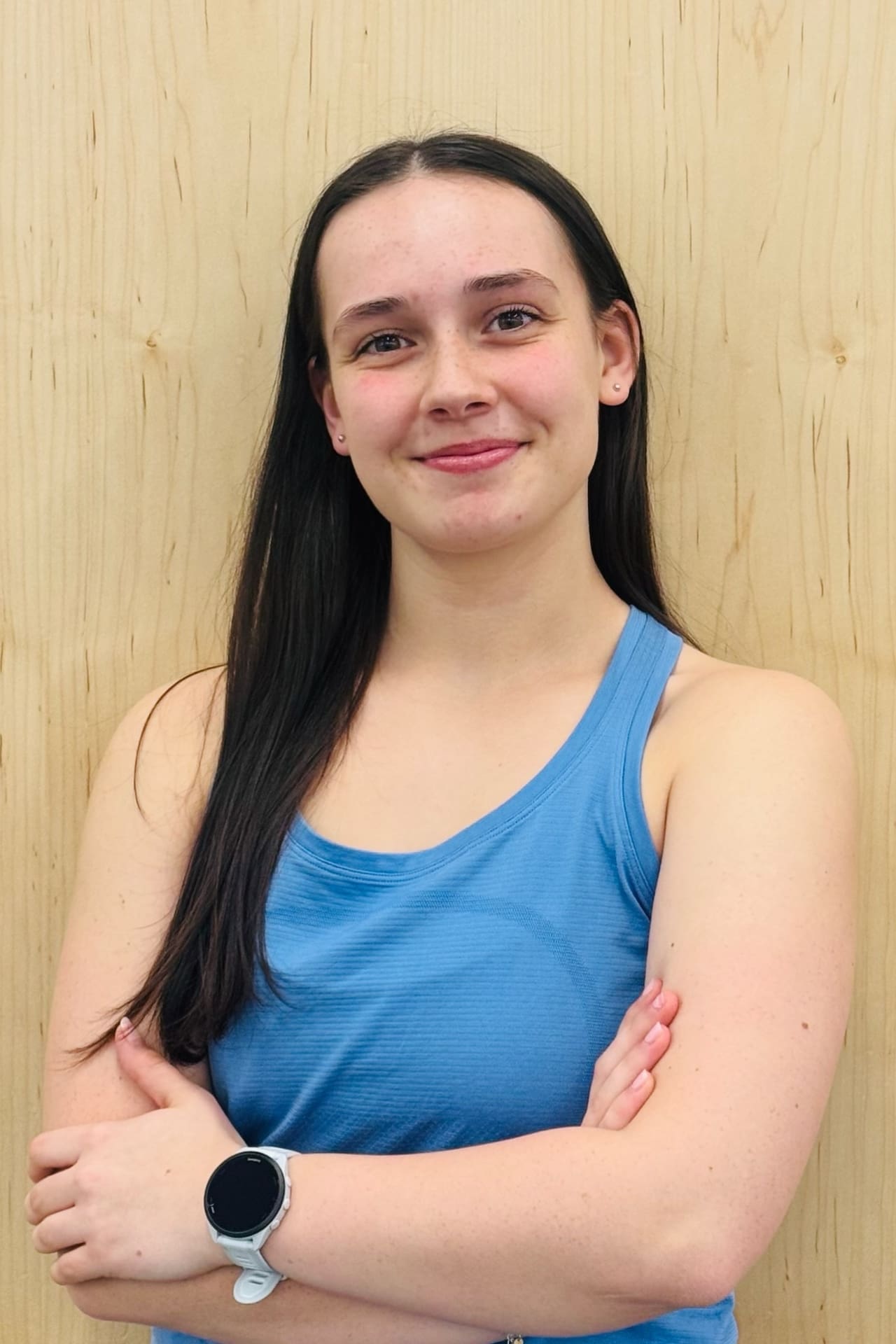 Young woman with long dark hair standing against a wooden wall, arms crossed, wearing a blue tank top and a smartwatch.