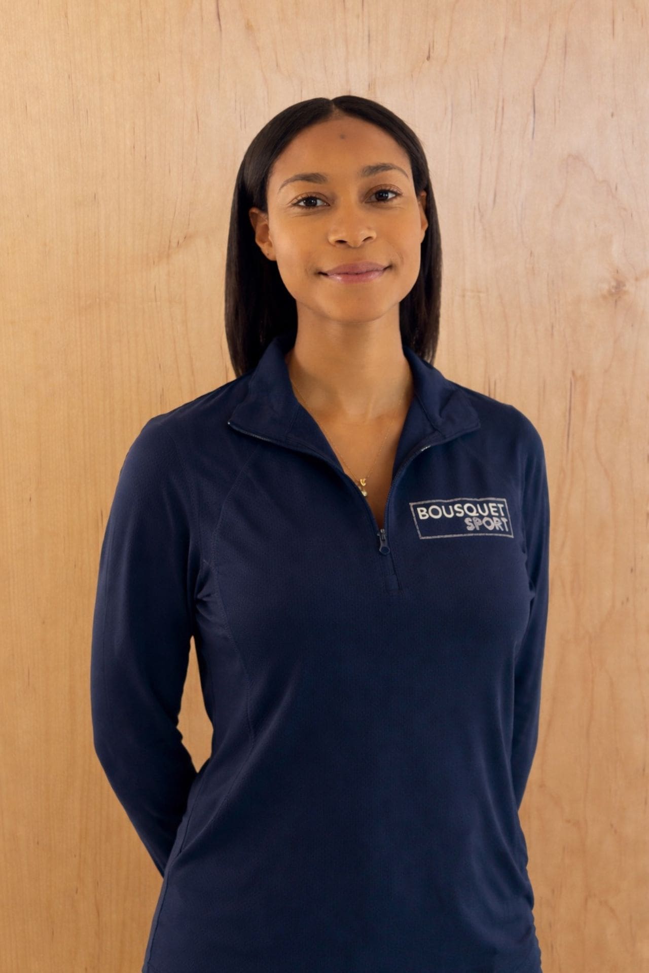 Portrait of a woman wearing a navy athletic top with a zipper and 'BOUSQUET SPORT' logo on the chest, standing against a wooden panel background.