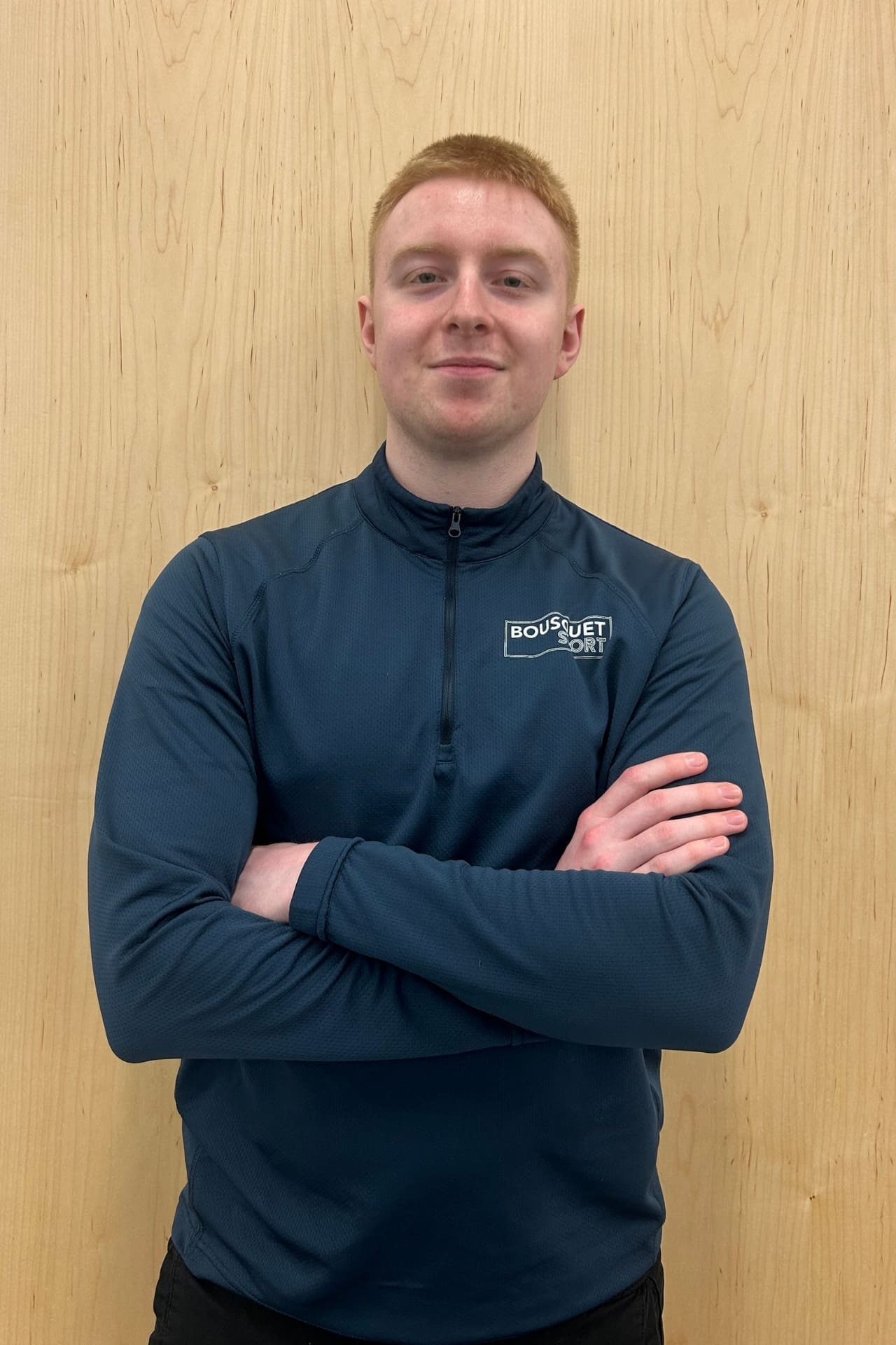 Young man standing against a wooden wall with arms crossed, wearing a dark blue athletic top with a small logo on the chest that reads BOUSQUET SPORT.