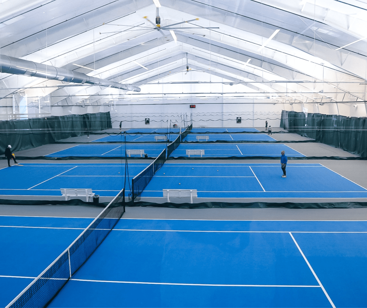 Indoor tennis facility with multiple blue courts, nets, and players practicing inside a large dome-like ceiling.