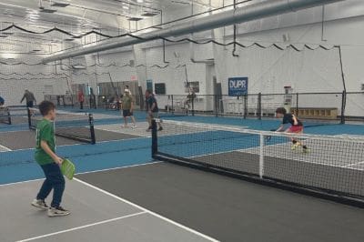 Indoor sports facility with multiple blue courts separated by nets; players on adjacent courts actively playing pickleball/tennis while others watch in the background.