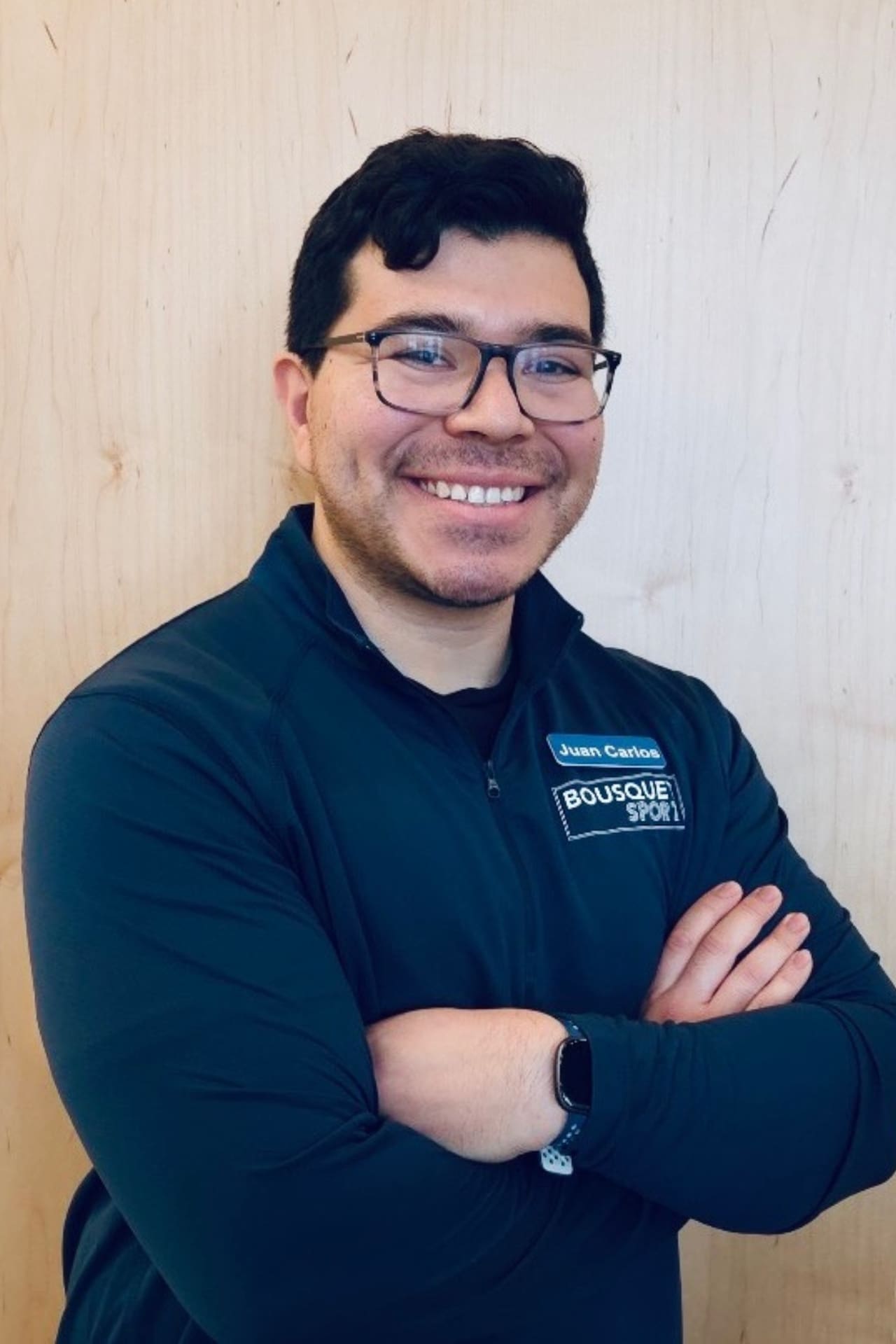 Smiling man with glasses and arms crossed, wearing a dark jacket with a Juan Carlos name badge on a light wood background.