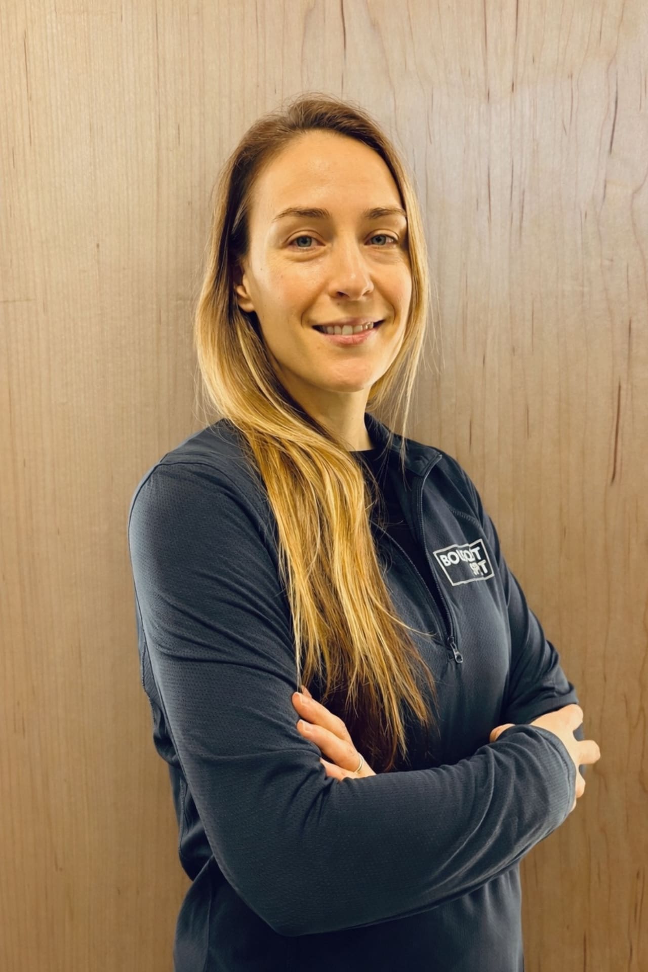 Smiling woman with long blonde-brown hair, arms crossed, wearing a dark athletic jacket against a wooden wall.