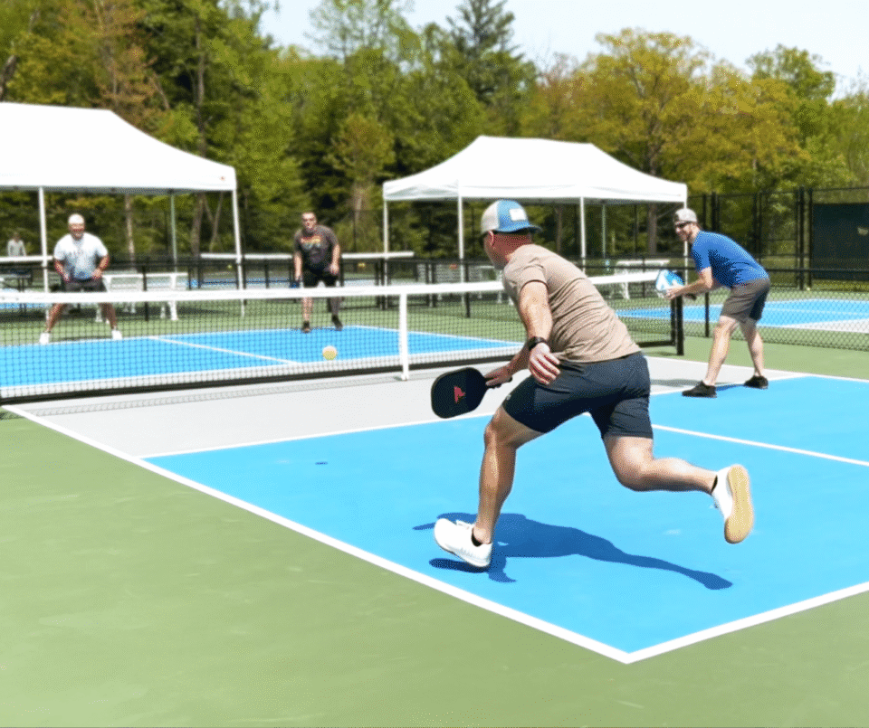 Three players compete in a pickleball match on a sunny outdoor court; the foreground player in a tan shirt and cap lunges with a paddle toward the ball.
