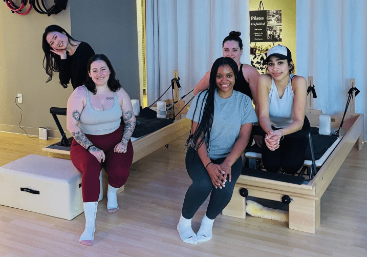 Five women in a Pilates studio pose for a group photo around reformer machines, smiling at the camera.
