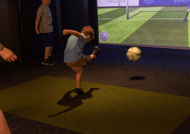 Young boy in a light blue shirt kicks a soccer ball on an indoor turf field, with a large screen projector displaying a goal in the background.