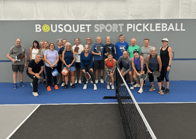 Group of adults posing with pickleball paddles on an indoor court in front of a wall reading 'BOUSQUET SPORT PICKLEBALL'.