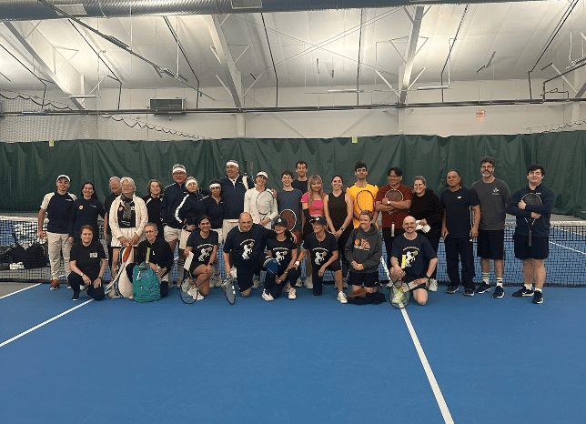 Group of people posing on an indoor tennis court with rackets, blue floor, and net in the background.
