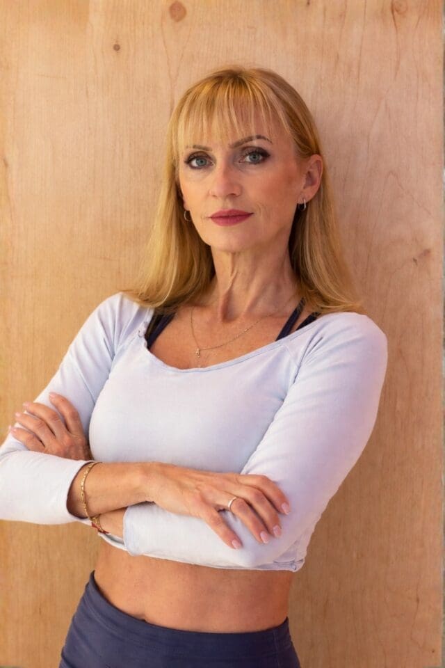 Confident woman with blonde hair in a light blue cropped top and dark leggings stands against a wooden wall with arms crossed.
